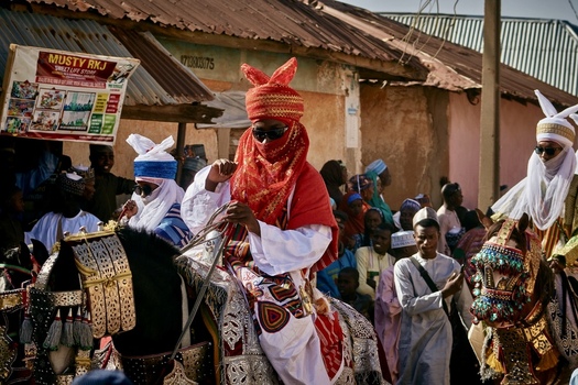Zaria City Nigeria 21 March 2026 View of a vibrant procession of horsemen in rich red and white regalia moving past earthy toned buildings under a bright sky. Zaria City Nigeria 21 March 2026 View of a vibrant procession of horsemen in rich red and white regalia moving past earthy toned buildings under a bright sky.