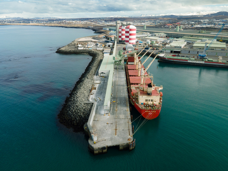 Hafnarfjordur Iceland 17 April 2026 Aerial view of a large red cargo ship docked at an industrial pier with red and white striped silos and snow capped mountains in the distance. Hafnarfjordur Iceland 17 April 2026 Aerial view of a large red cargo ship docked at an industrial pier with red and white striped silos and snow capped mountains in the distance.
