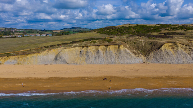 Aerial view of the dramatic sandstone cliffs and golden sandy beach at West Bay with people walking along the shoreline under a blue sky in West Bay England United Kingdom. Aerial view of the dramatic sandstone cliffs and golden sandy beach at West Bay with people walking along the shoreline under a blue sky in West Bay England United Kingdom.
