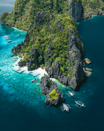 Aerial view of dramatic limestone cliffs and turquoise waters with traditional outrigger boats anchored near the white sand beach in El Nido Mimaropa Philippines. Aerial view of dramatic limestone cliffs and turquoise waters with traditional outrigger boats anchored near the white sand beach in El Nido Mimaropa Philippines.