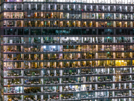 Aerial view of illuminated office windows in a modern glass skyscraper at night showing desks plants and people working in Poland. Aerial view of illuminated office windows in a modern glass skyscraper at night showing desks plants and people working in Poland.
