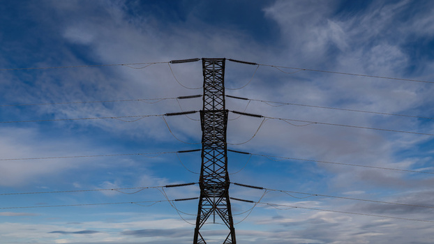 Aerial view of a high voltage power pylon with steel lattice structure and electrical lines stretching across a cloudy blue sky in Iceland.