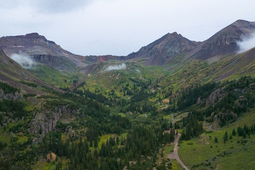 Aerial view of Mt. Sneffels and the lush green landscape of Yankee Boy Basin with pine forests rugged mountain peaks and a dirt trail in Ouray Colorado United States.