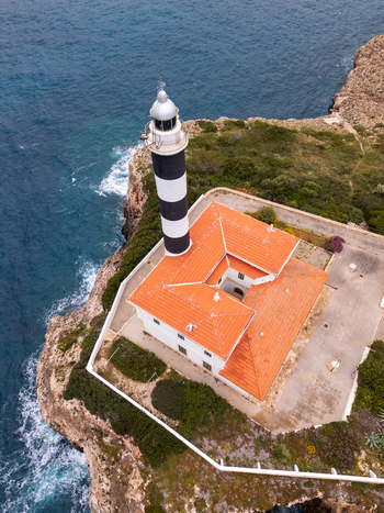 Aerial view of the Portocolom Lighthouse with its black and white stripes and red tiled building on a rocky cliff by the sea in Portocolom Balearic Islands Spain. Aerial view of the Portocolom Lighthouse with its black and white stripes and red tiled building on a rocky cliff by the sea in Portocolom Balearic Islands Spain.