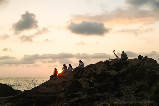 Sri Lanka 25 January 2026 View of people sitting on a rocky cliff overlooking the ocean during a golden sunset with soft clouds.
