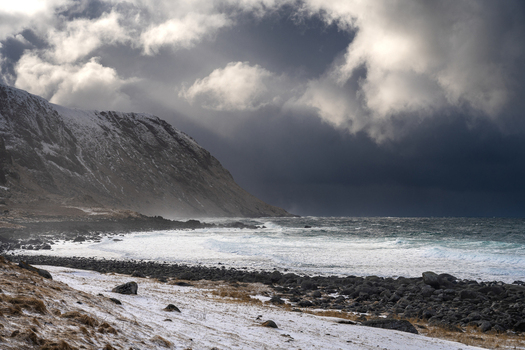 View of a snowy coastal landscape with a rocky beach crashing waves and dramatic dark storm clouds over the mountains in Leknes Lofoten Norway.
