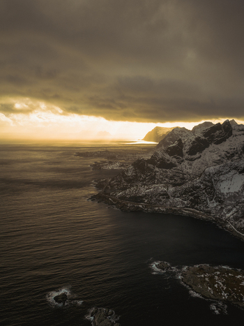 Aerial view of rugged snow dusted mountains meet the dark turbulent sea under a brooding sky Reinebringen Nordland Norway.