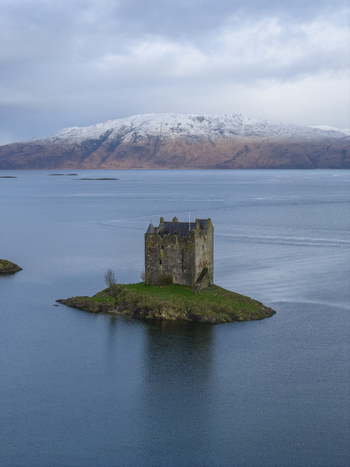 Aerial view of Castle Stalker on a small tidal islet surrounded by the calm waters of Loch Laich with snow capped mountains under a cloudy sky Appin Scotland United Kingdom. Aerial view of Castle Stalker on a small tidal islet surrounded by the calm waters of Loch Laich with snow capped mountains under a cloudy sky Appin Scotland United Kingdom.