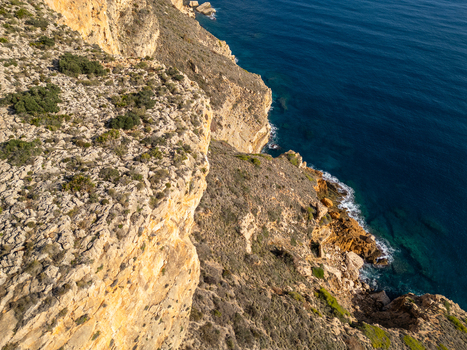 Aerial view of Cala Llebeig featuring rugged rocky cliffs dropping into the deep blue Mediterranean Sea with sparse coastal vegetation in El Poble Nou de Benitatxell Valencian Community Spain.