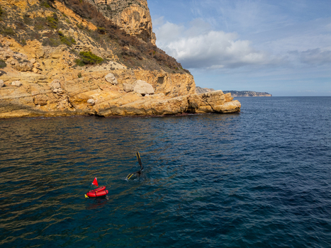 Aerial view of a diver with a red surface marker buoy in the clear blue waters of Cala Llebeig near rocky cliffs in El Poble Nou de Benitatxell Valencian Community Spain.