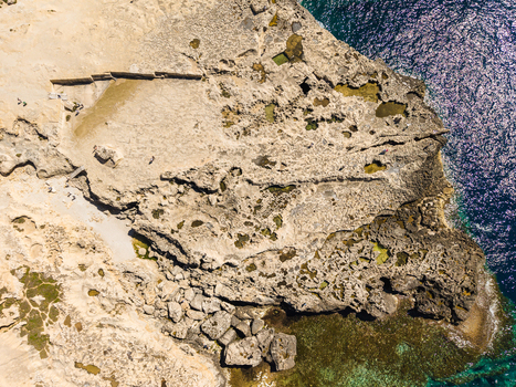 Aerial view of the rugged limestone coastline and natural rock formations at Dwejra Bay with clear turquoise water and people walking on the rocks Gozo Region Malta. Aerial view of the rugged limestone coastline and natural rock formations at Dwejra Bay with clear turquoise water and people walking on the rocks Gozo Region Malta.