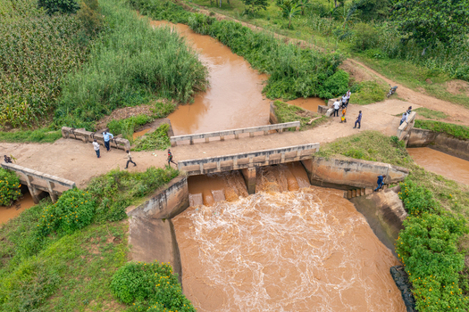 Aerial view of a concrete bridge over a muddy river with people standing on the structure surrounded by lush vegetation and agricultural fields in Butaleja Eastern Region Uganda. Aerial view of a concrete bridge over a muddy river with people standing on the structure surrounded by lush vegetation and agricultural fields in Butaleja Eastern Region Uganda.