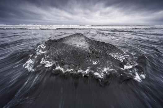 View of a large iceberg fragment on the black sand with rushing ocean water under a dramatic cloudy sky at Diamond Beach Sveitarfelagid Hornafjordur Iceland.