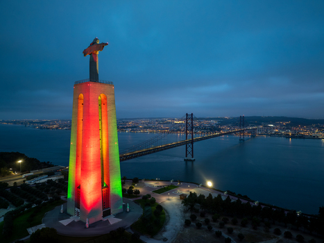Aerial view of the Sanctuary of Christ the King illuminated in red and green with the 25 de Abril Bridge and Tagus River at dusk in Almada Setubal Portugal. Aerial view of the Sanctuary of Christ the King illuminated in red and green with the 25 de Abril Bridge and Tagus River at dusk in Almada Setubal Portugal.
