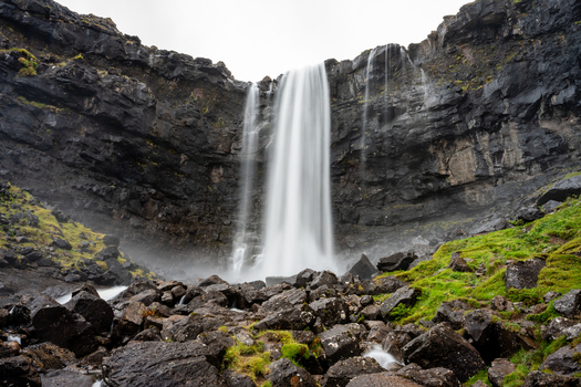 View of a powerful waterfall cascading over dark basalt cliffs with mossy green slopes and scattered rocks in the foreground Faroe Islands.