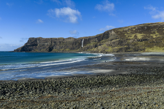 Aerial view of Talisker Bay Beach with its dark volcanic sand turquoise waves and a waterfall cascading down the rugged cliffs under a clear blue sky Isle of Skye Scotland United Kingdom. Aerial view of Talisker Bay Beach with its dark volcanic sand turquoise waves and a waterfall cascading down the rugged cliffs under a clear blue sky Isle of Skye Scotland United Kingdom.