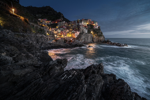View of the colorful houses of Manarola village perched on a cliff above the rocky coastline and crashing waves at twilight in Manarola Liguria Italy. View of the colorful houses of Manarola village perched on a cliff above the rocky coastline and crashing waves at twilight in Manarola Liguria Italy.