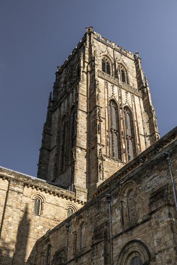 View of the historic stone tower of Durham Cathedral featuring intricate Gothic architecture arched windows and weathered masonry under a clear blue sky in Durham England United Kingdom. View of the historic stone tower of Durham Cathedral featuring intricate Gothic architecture arched windows and weathered masonry under a clear blue sky in Durham England United Kingdom.