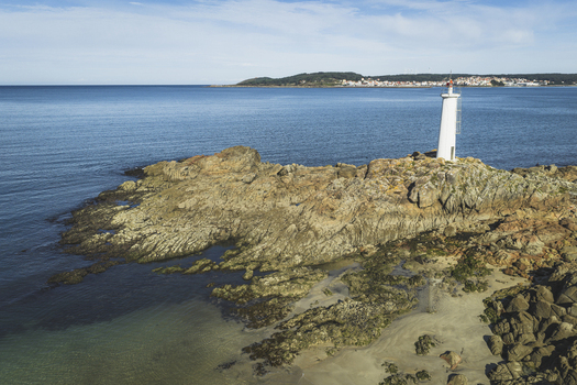Aerial view of a white lighthouse standing on rugged rocky cliffs overlooking the calm blue sea with a distant coastline in Galicia Spain.