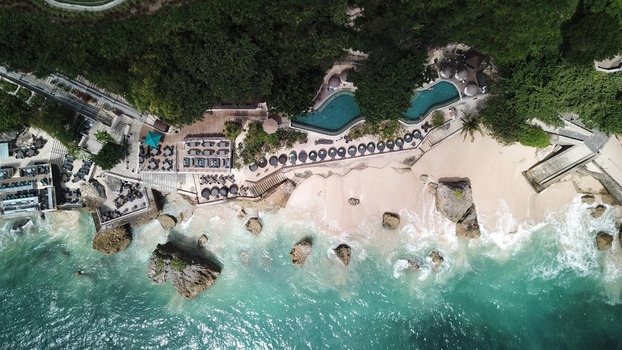 Indonesia 20 February 2026 Aerial view of a coastal resort featuring a swimming pool lounge chairs on a sandy beach turquoise water and large rocks along the shoreline.