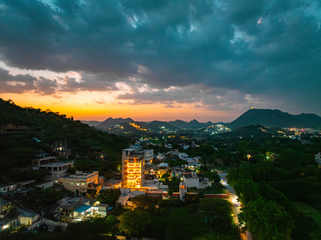 Aerial view of illuminated buildings nestled among lush greenery and rolling hills under a dramatic sunset sky in Udaipur Rajasthan India.