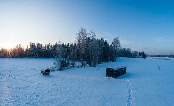 Aerial view of a barrel sauna and a modern cabin situated in a vast snow covered field near a dense forest during a clear winter sunset in Latvia. Aerial view of a barrel sauna and a modern cabin situated in a vast snow covered field near a dense forest during a clear winter sunset in Latvia.