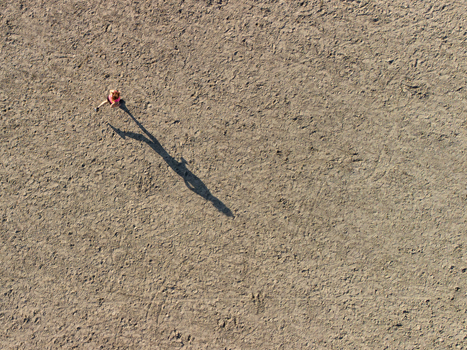 Aerial view of a woman in pink walking across a sandy surface with a long shadow cast on the ground in Almere Flevoland Netherlands.