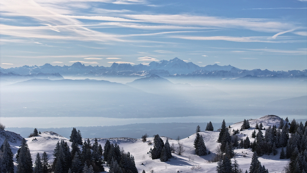 Aerial view of snow dusted evergreens meet the misty valley and hazy mountain range under a striated sky. Gex Auvergne Rhône Alpes France. Aerial view of snow dusted evergreens meet the misty valley and hazy mountain range under a striated sky. Gex Auvergne Rhône Alpes France.