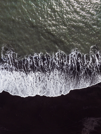 Aerial view of white sea foam crashing onto the dark black sand beach with rhythmic waves and contrast between the dark volcanic sand and the Atlantic Ocean in Iceland. Aerial view of white sea foam crashing onto the dark black sand beach with rhythmic waves and contrast between the dark volcanic sand and the Atlantic Ocean in Iceland.