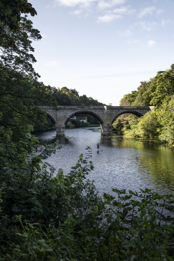 View of a person on a stand up paddleboard on the River Wear passing under a stone arch bridge surrounded by lush green trees in Durham England United Kingdom. View of a person on a stand up paddleboard on the River Wear passing under a stone arch bridge surrounded by lush green trees in Durham England United Kingdom.