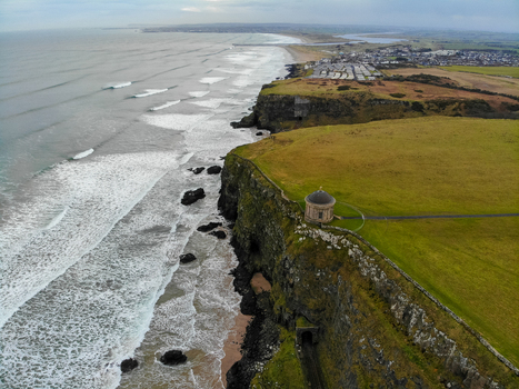 Aerial view of Mussenden Temple on dramatic cliffs overlooking the Atlantic Ocean with crashing waves on the beach and green fields in Coleraine Northern Ireland United Kingdom. Aerial view of Mussenden Temple on dramatic cliffs overlooking the Atlantic Ocean with crashing waves on the beach and green fields in Coleraine Northern Ireland United Kingdom.