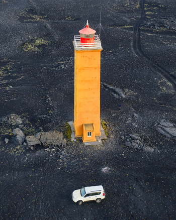 Aerial view of a yellow lighthouse with a red lantern room standing on a vast black volcanic field with a white SUV parked nearby in Kirkjubaejarklaustur Skaftarhreppur Iceland. Aerial view of a yellow lighthouse with a red lantern room standing on a vast black volcanic field with a white SUV parked nearby in Kirkjubaejarklaustur Skaftarhreppur Iceland.