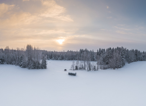 Aerial view of a snow covered field with a small structure near a forest edge under a soft winter sky in Latvia. Aerial view of a snow covered field with a small structure near a forest edge under a soft winter sky in Latvia.