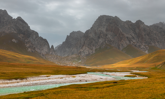 View of the turquoise river flowing through the grassy plains of Kol Suu framed by rugged mountain peaks under a cloudy sky in At Bashi District Naryn Region Kyrgyzstan. View of the turquoise river flowing through the grassy plains of Kol Suu framed by rugged mountain peaks under a cloudy sky in At Bashi District Naryn Region Kyrgyzstan.