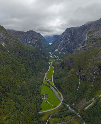 Aerial view of a winding road and river flowing through a deep valley surrounded by steep mountains and green fields in Gudvangen Vestland Norway.