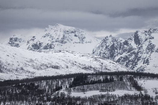 View of snow covered mountain peaks and a dark forest under a cloudy sky in Tromso Troms Norway.