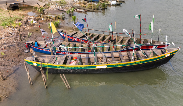 Aerial view of colorful wooden boats moored along the muddy riverbank with flags and people visible in Minima Rivers Nigeria.