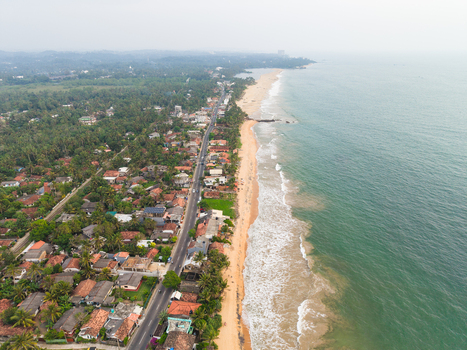 Aerial view of the coastline featuring a sandy beach turquoise ocean waves and a coastal road lined with houses and palm trees in Galle Southern Province Sri Lanka.