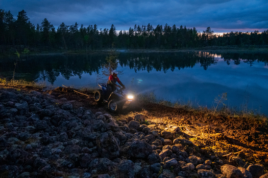View of a person riding an ATV along the rocky muddy shore of Lake Ladoga at twilight with forest reflections in the water in Sortavala Respublika Kareliya Russia.