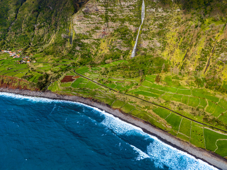 Aerial view of a dramatic coastline where the rugged verdant cliffs meet the deep blue sea with a waterfall cascading down the slopes Faj&atilde; Grande Azores Portugal.