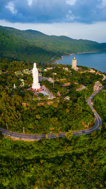 Aerial view of the Lady Buddha statue at Linh Ung Pagoda surrounded by lush green hills a winding coastal road and the blue sea in Da Nang Vietnam. Aerial view of the Lady Buddha statue at Linh Ung Pagoda surrounded by lush green hills a winding coastal road and the blue sea in Da Nang Vietnam.