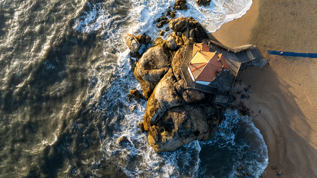 Aerial view of Capela do Senhor da Pedra built on a rocky outcrop with ocean waves crashing against the shore at Miramar Porto Portugal.