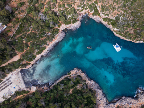 Aerial view of Cala Sa Nau cove with turquoise water anchored boats and rocky cliffs surrounded by green trees in Felanitx Balearic Islands Spain. Aerial view of Cala Sa Nau cove with turquoise water anchored boats and rocky cliffs surrounded by green trees in Felanitx Balearic Islands Spain.