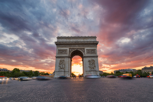 View of the Arc de Triomphe under a dramatic sunset sky with purple clouds and motion blurred traffic on the cobblestone street in Paris Ile de France France.