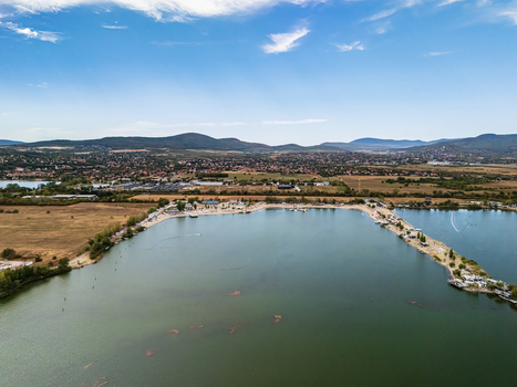 Aerial view of Lake Lupa s beach and recreational waterfront in Budakalasz Hungary with open water rural fields a nearby town and the Buda Hills beyond.