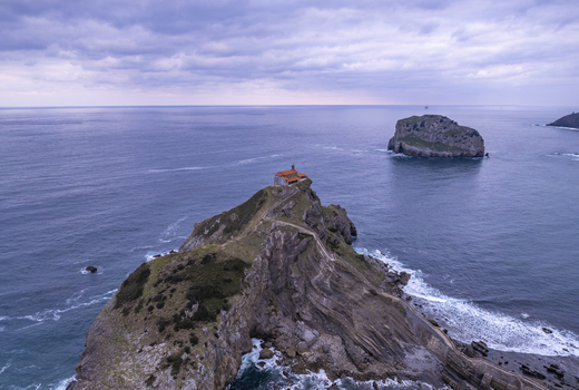 Aerial view of San Juan de Gaztelugatxe perched on a rocky islet connected by a winding stone bridge surrounded by the deep blue waters of the Bay of Biscay under a cloudy sky Gaztelugatxeko Doniene Euskadi Spain.