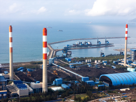 Aerial view of a coal fired power plant with tall red and white chimneys coal storage piles industrial buildings and a harbor along the coastline in Cilacap Central Java Indonesia. Aerial view of a coal fired power plant with tall red and white chimneys coal storage piles industrial buildings and a harbor along the coastline in Cilacap Central Java Indonesia.
