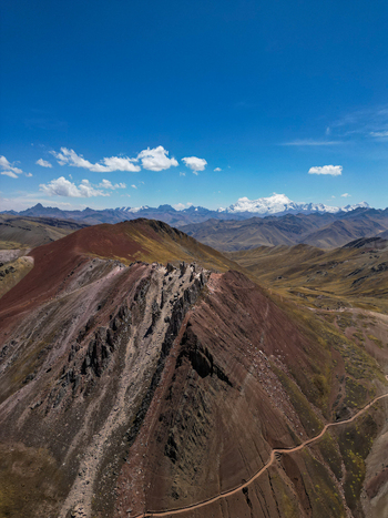 Aerial view of Vinicunca mountain with its reddish slopes and rocky ridge overlooking a winding trail and distant snow capped peaks in Quispicanchi Province Cusco Peru.