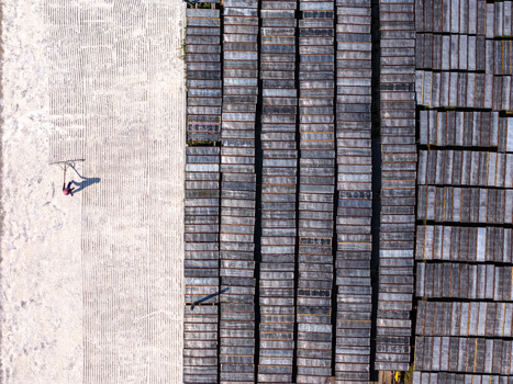 Aerial view of a worker raking material on a large light colored surface next to rows of dark rectangular frames in Polanharjo Central Java Indonesia.