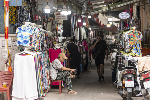 Hanoi Ha Noi Vietnam 11 November 2025 View of a bustling narrow street market in the Old Quarter with colorful clothing racks hanging lights motorbikes and people walking.